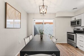 A modern kitchen with a dining table, chairs, a hanging light fixture, and a view through a glass door.