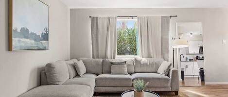 A modern living room with a sectional sofa, a round coffee table with a plant, and a view of a kitchen and outdoor greenery through large windows.