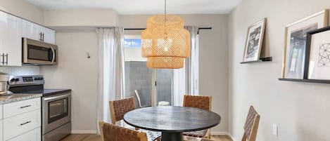A modern kitchen with a dining area, featuring a round table and wicker chairs, a hanging light fixture, and a microwave.