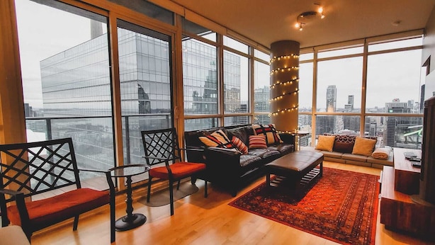 A modern living room with a leather sofa, red chairs, a coffee table, and a large window offering a city view.