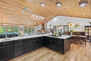 A modern kitchen with black cabinets, a large island, and a dining area with wooden chairs. - Luxe Lake Home | Hot Tub + Private Dock & Beach (Nine Mile Falls)