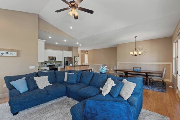 A living room with a sectional sofa, a ceiling fan, a dining area with a table and chairs, and a kitchen in the background.