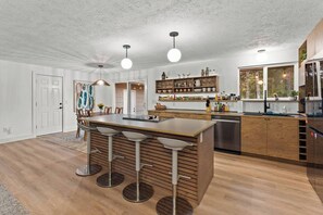 A modern kitchen with a central island, bar stools, pendant lighting, and wooden cabinetry. - Game On | 2 Lounges, Ping Pong, Firepit + Hot Tub (Spokane)