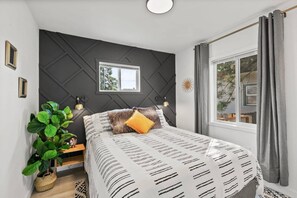 A modern bedroom with a bed, a window, a plant, and a wall-mounted light fixture. - Family- & pet-friendly Spokane home: 3BR/2BA, fenced yard, near downtown. (Spokane)