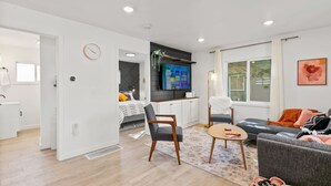 Una sala de estar moderna con un sofá, una silla, un televisor montado en la pared y una zona de comedor con mesa y sillas.