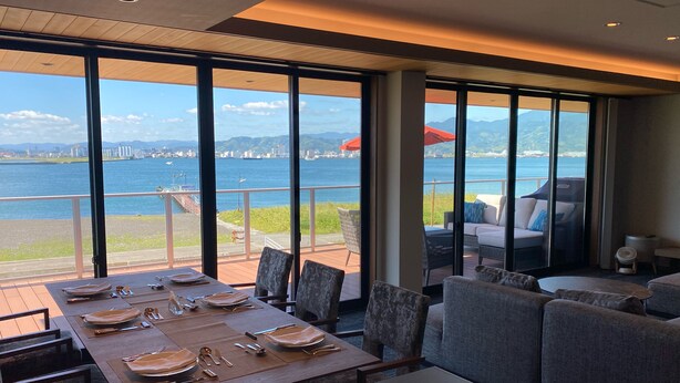 A dining area with a table set for four, overlooking a waterfront with a cityscape in the background. - 'Ulalena (Shizuoka)