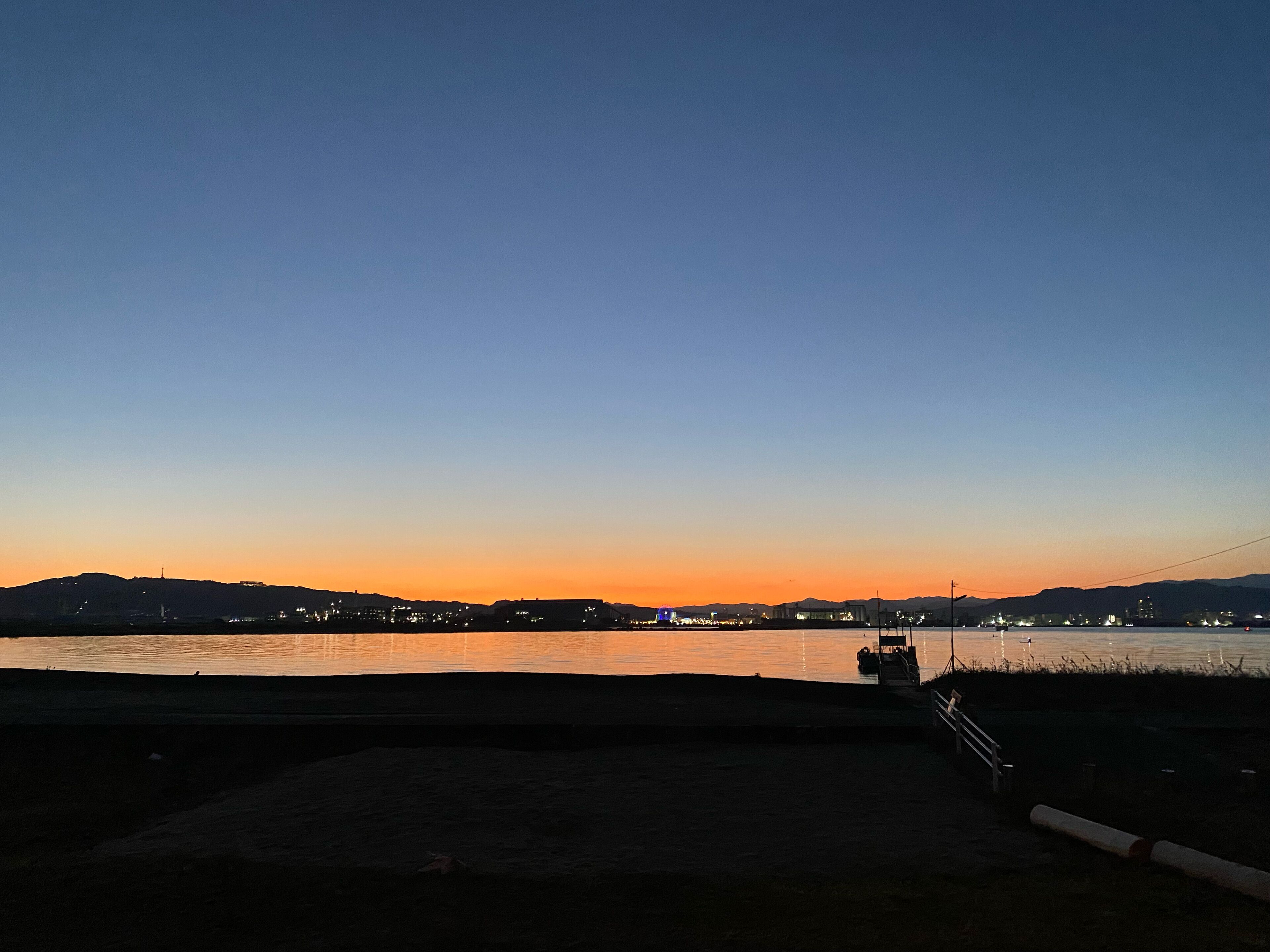 A sunset view over a body of water with a dock and distant city lights.