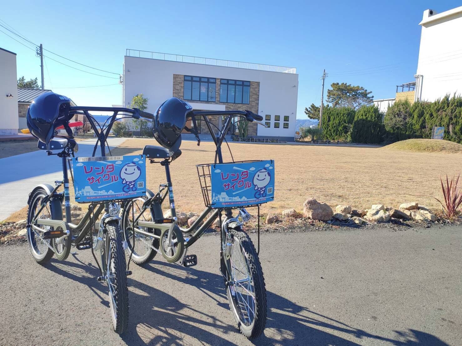 Dos bicicletas con cascos y carteles azules adheridos que muestran un personaje de dibujos animados, estacionadas en un estacionamiento con un edificio al fondo.