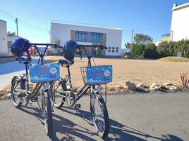 Dos bicicletas con cascos y carteles azules adheridos que muestran un personaje de dibujos animados, estacionadas en un estacionamiento con un edificio al fondo.