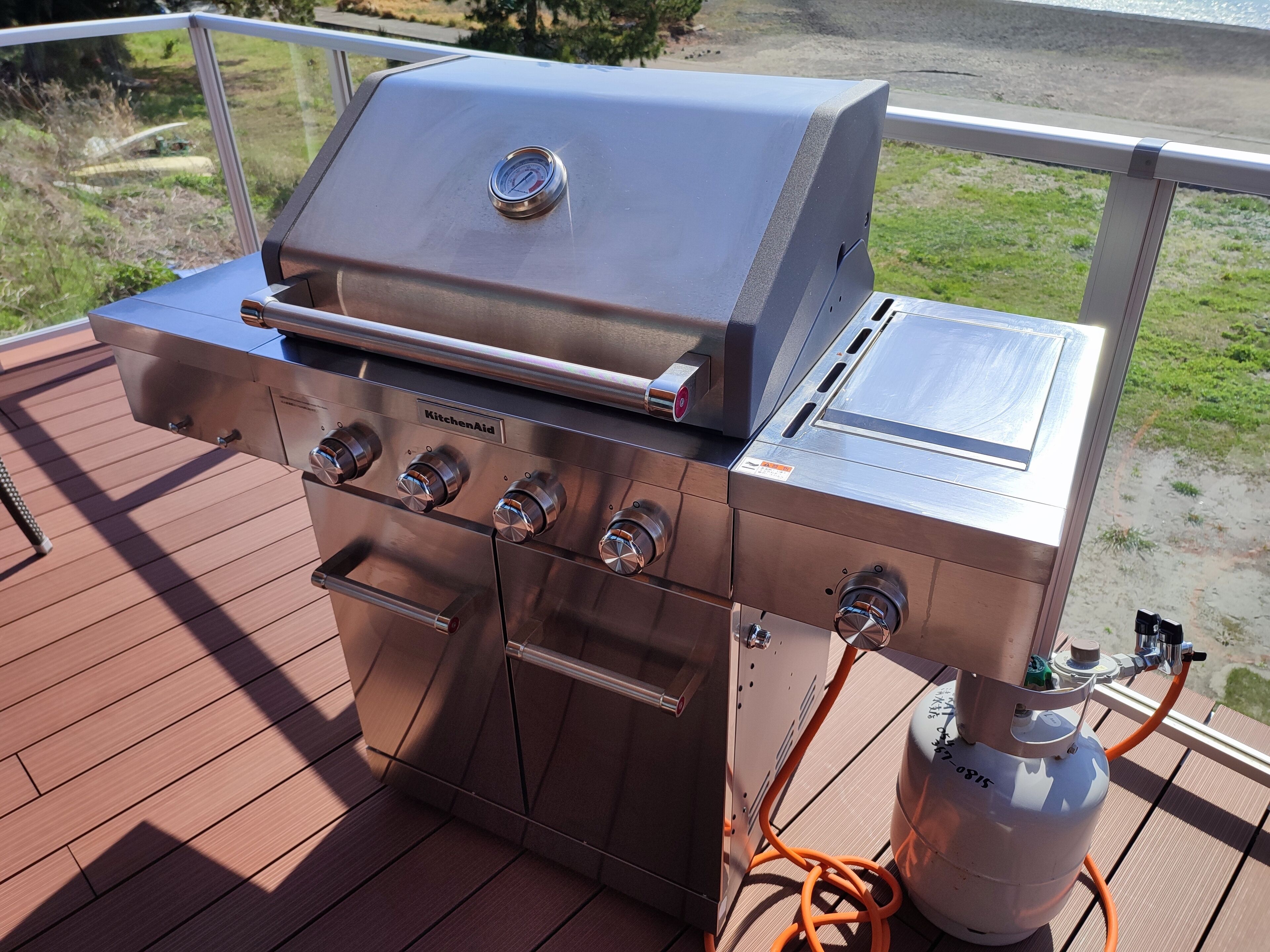 Een roestvrijstalen buitenbarbecue met vier branders en een ingebouwde thermometer, aangesloten op een propaantank.