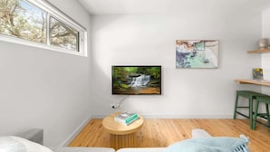 Un salon moderne avec un grand téléviseur fixé au mur, une table basse en bois sur laquelle sont posés des livres, un fauteuil vert et une vue sur les arbres par une grande fenêtre.