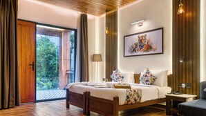 A spacious bedroom with a large bed, a wooden headboard, a framed tiger artwork, and a view of greenery through a sliding door. - Corbett Machaan Resort (Ramnagar)