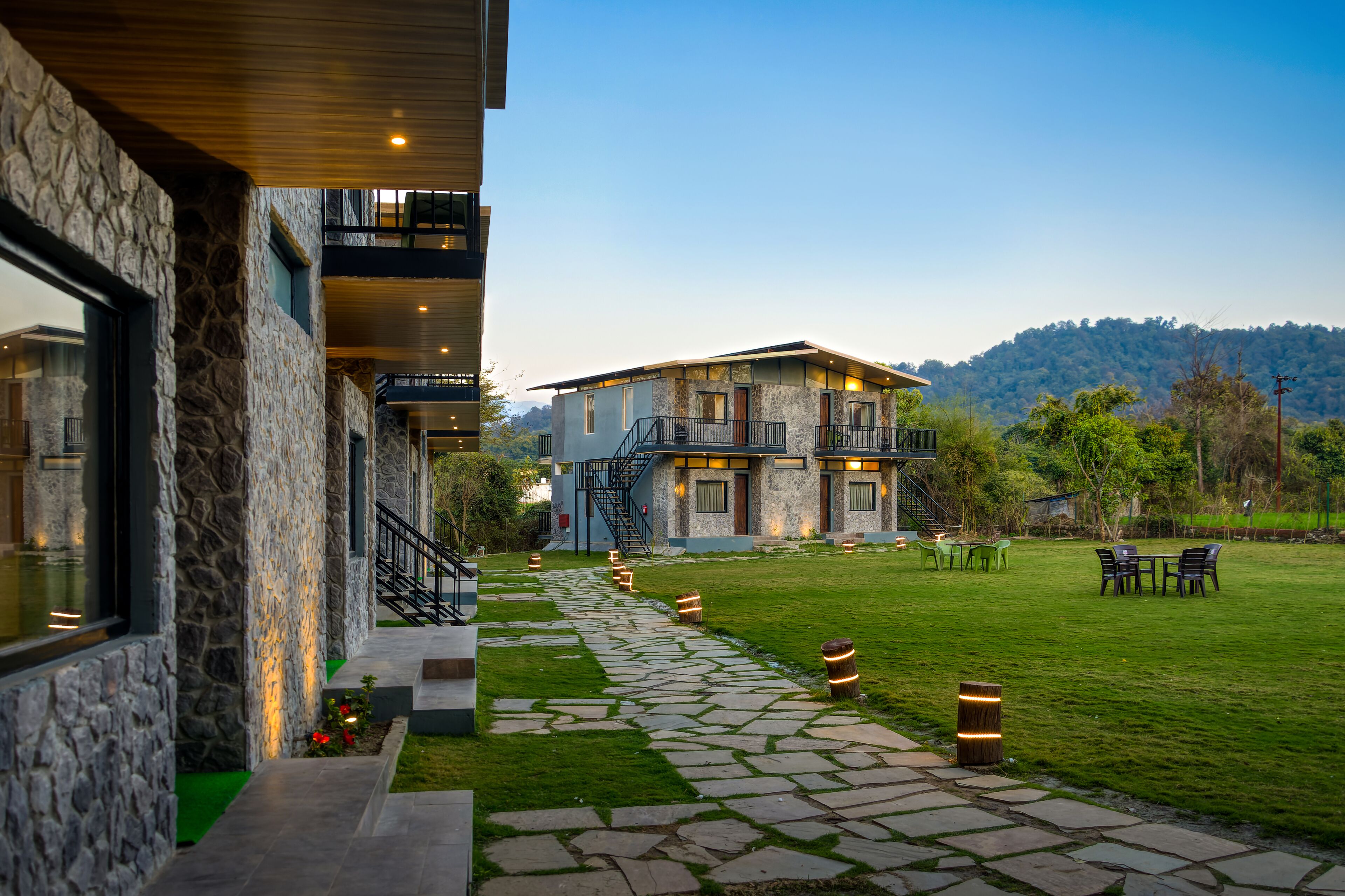 A modern building with a stone facade, a stone pathway leading to a grassy area with outdoor seating, and a staircase to an upper level.