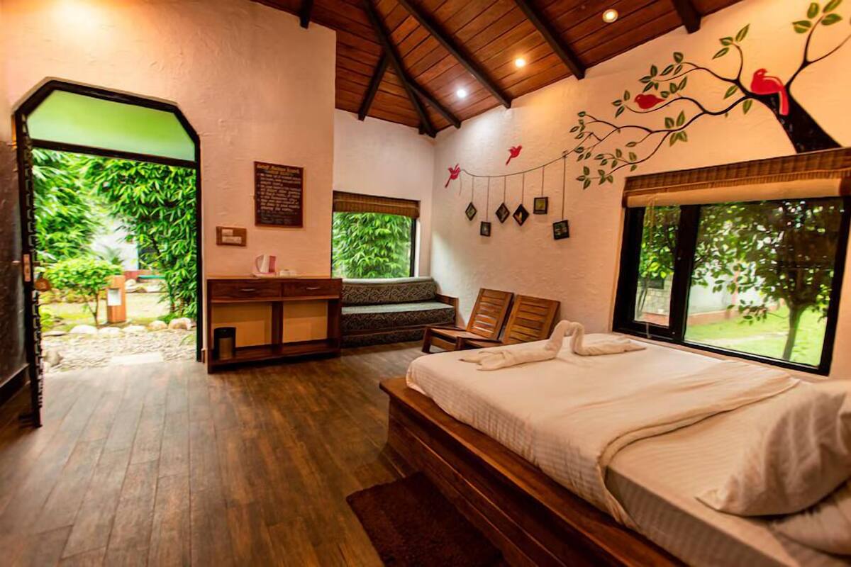 A bedroom with a large bed, a wooden bench, a small table, and a view of greenery through a window.