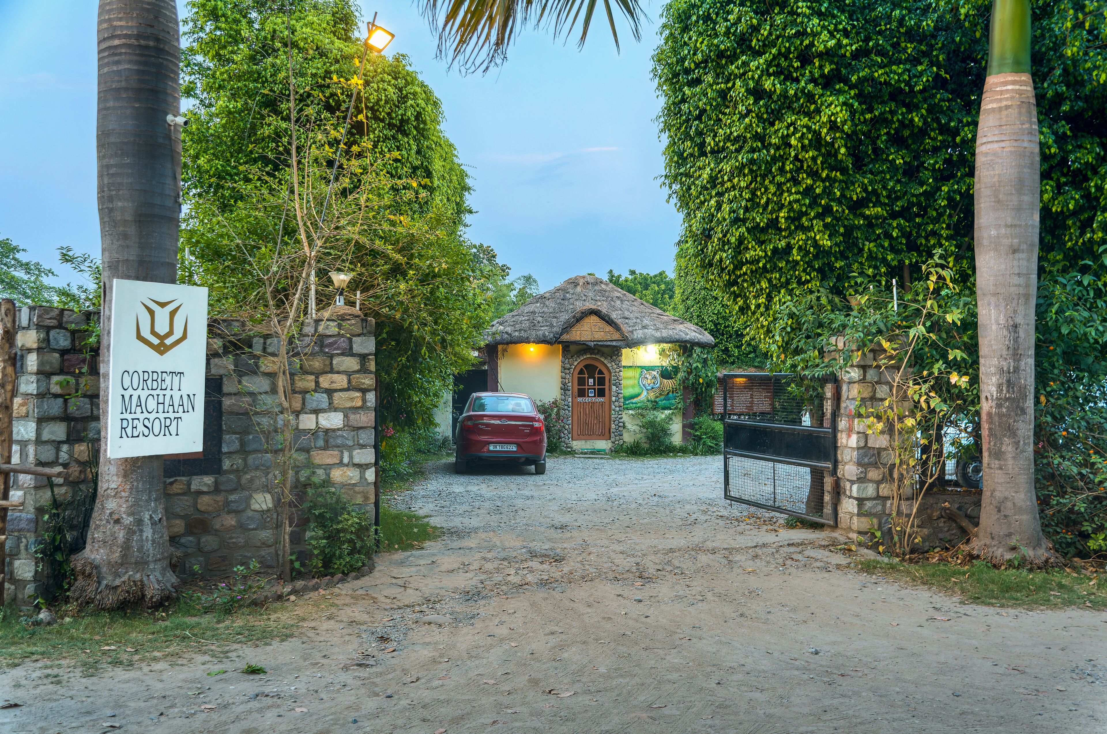 A rustic entrance to Corbett Machaan Resort with a thatched-roof structure, surrounded by lush greenery and a stone wall.