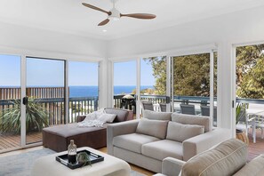 Amplia sala de estar con un ventanal que ofrece vista al mar, un ventilador de techo y una terraza de madera afuera.