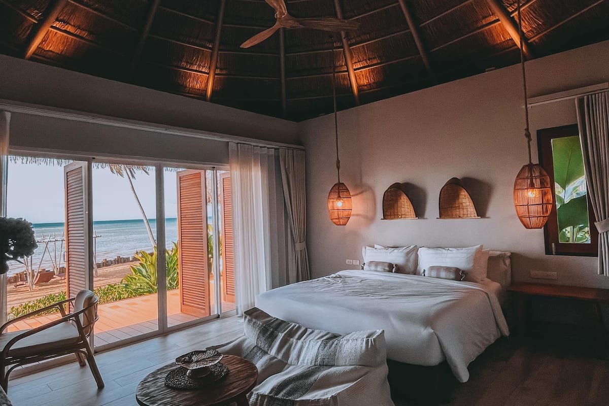 A bedroom with a bed, a chair, a table, and a view of the beach.
