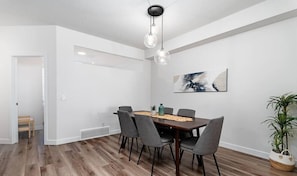 Uma área de jantar moderna com uma mesa de madeira, cadeiras estofadas cinzentas, um lustre pendente e uma pintura abstrata na parede.