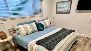 A bedroom with a bed, a nightstand, a lamp, a window with blinds, a framed picture on the wall, and a television. - 10 Steps to the Sand…Capitola Beach Front at the mouth of the Wharf-Pet Friendly (Capitola)