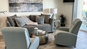 A modern living room with a grey sofa set, a round coffee table, and a grey armchair. A large abstract painting is on the wall, and a flat-screen TV is mounted above a sliding glass door. - 10 Steps to the Sand…Capitola Beach Front at the mouth of the Wharf-Pet Friendly (Capitola)