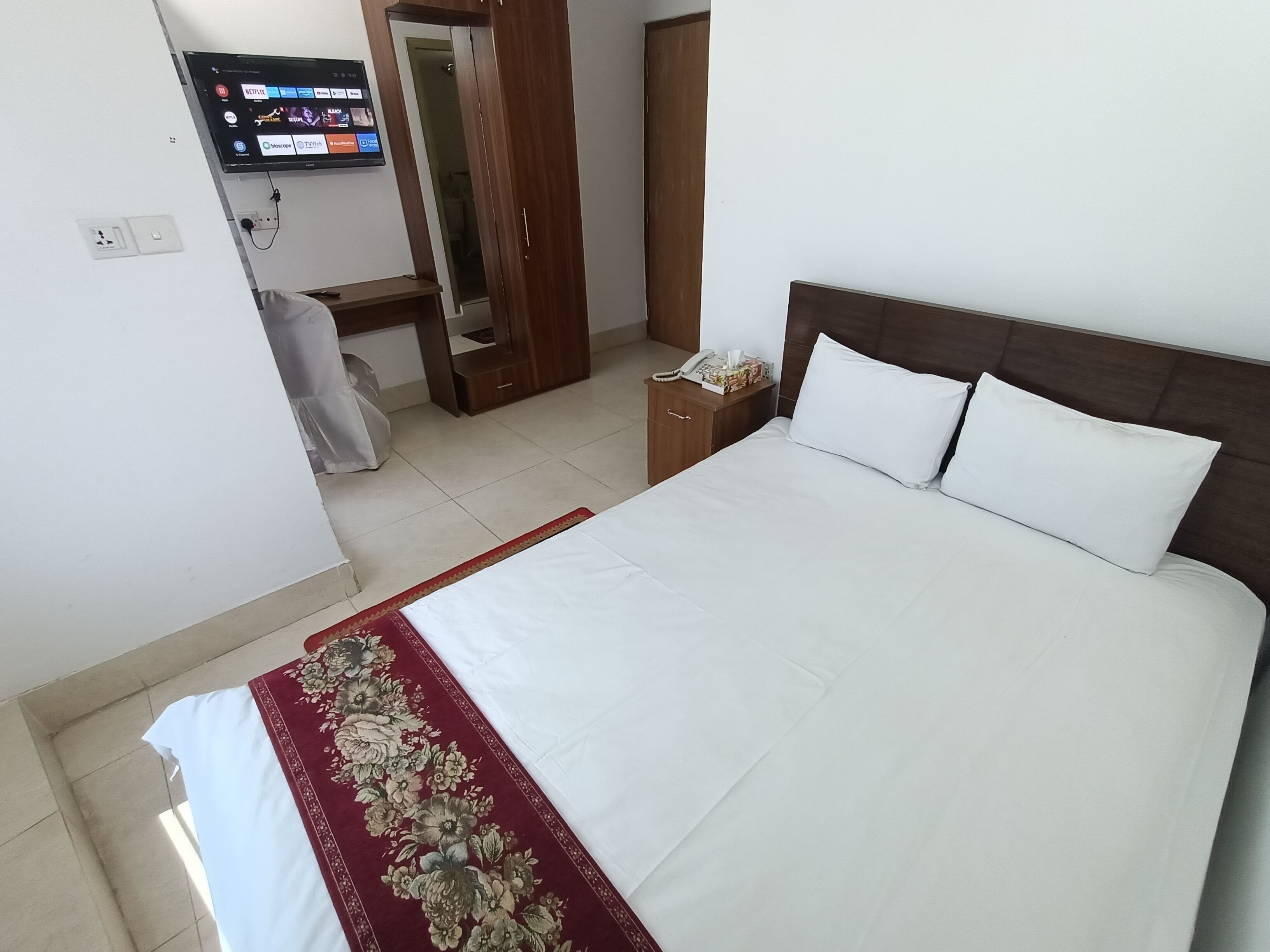 Habitación de hotel con una cama grande, un televisor montado en la pared, un armario de madera y una mesita de noche con un teléfono y pañuelos.