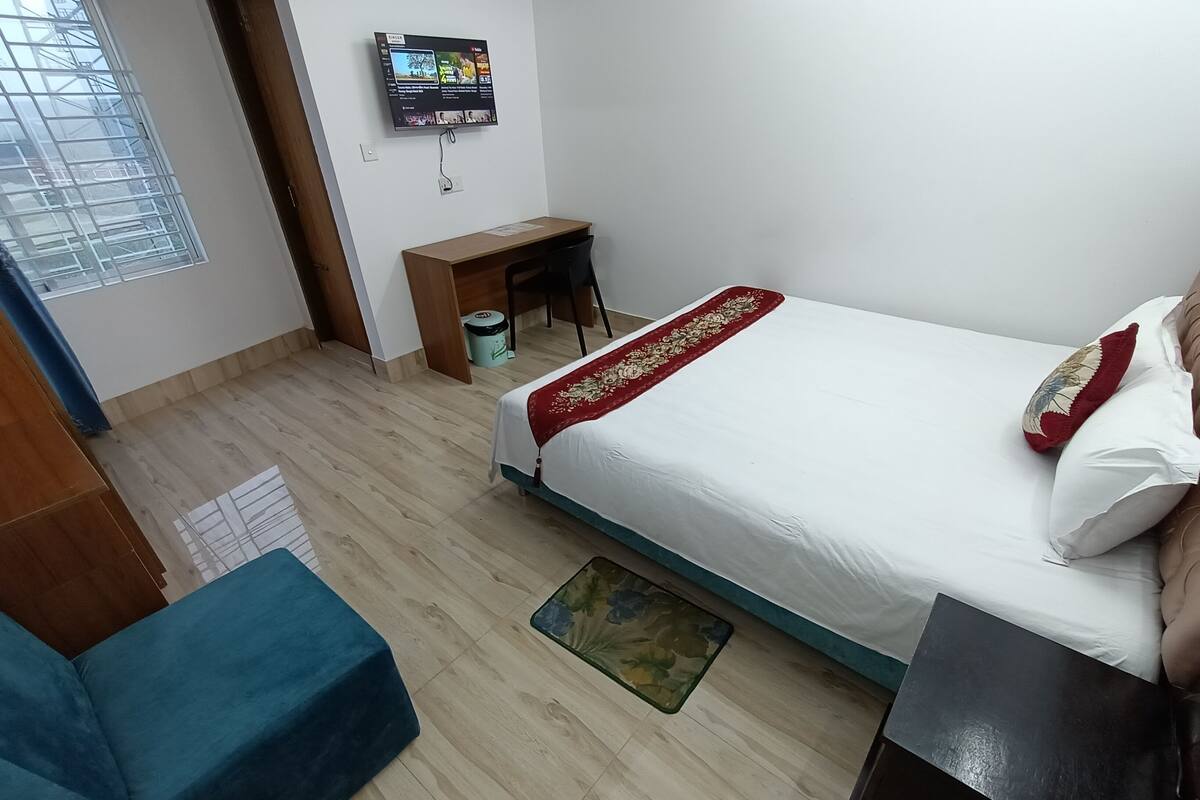 A hotel room with a bed, a desk, a TV mounted on the wall, and a window with blinds.