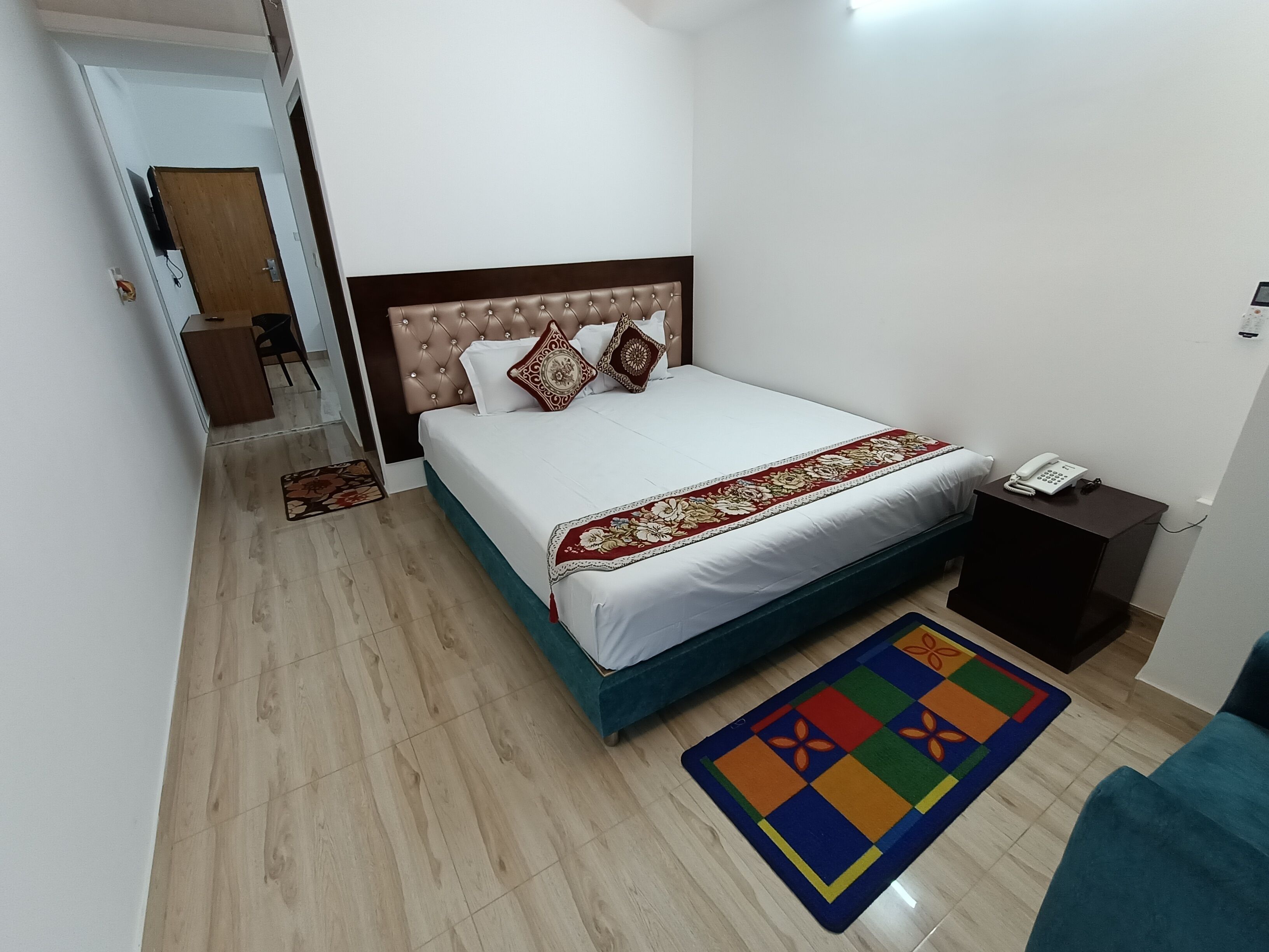 A bedroom with a bed, a bedside table with a phone, a colorful rug, and a wooden floor.