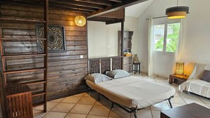 A bedroom with a wooden wall, a bed, a nightstand, a lamp, and a window with a view of greenery. - Ylang6 Avec vue Panoramique (Saint Leu)