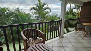 A balcony with a wooden chair and a table, surrounded by a metal railing, overlooking a lush garden with palm trees. - Ylang6 Avec vue Panoramique (Saint Leu)