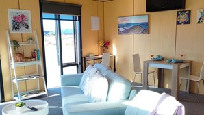 A modern living room with a light blue sofa, a white side table, and a small round table. There is a bookshelf with decorative items, a flat-screen TV mounted on the wall, and a dining area with a table and chairs.