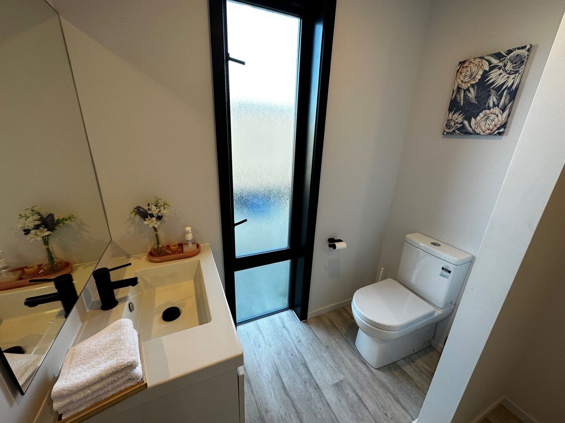 A modern bathroom with a white sink, mirror, and toilet. There is a window with frosted glass, a towel rack, and a floral artwork on the wall.