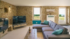 Ein modernes Wohnzimmer mit Steinwänden, einem Flachbildfernseher, einem grauen Sofa mit türkisfarbenen Kissen und einem großen Fenster mit Blick ins Freie.