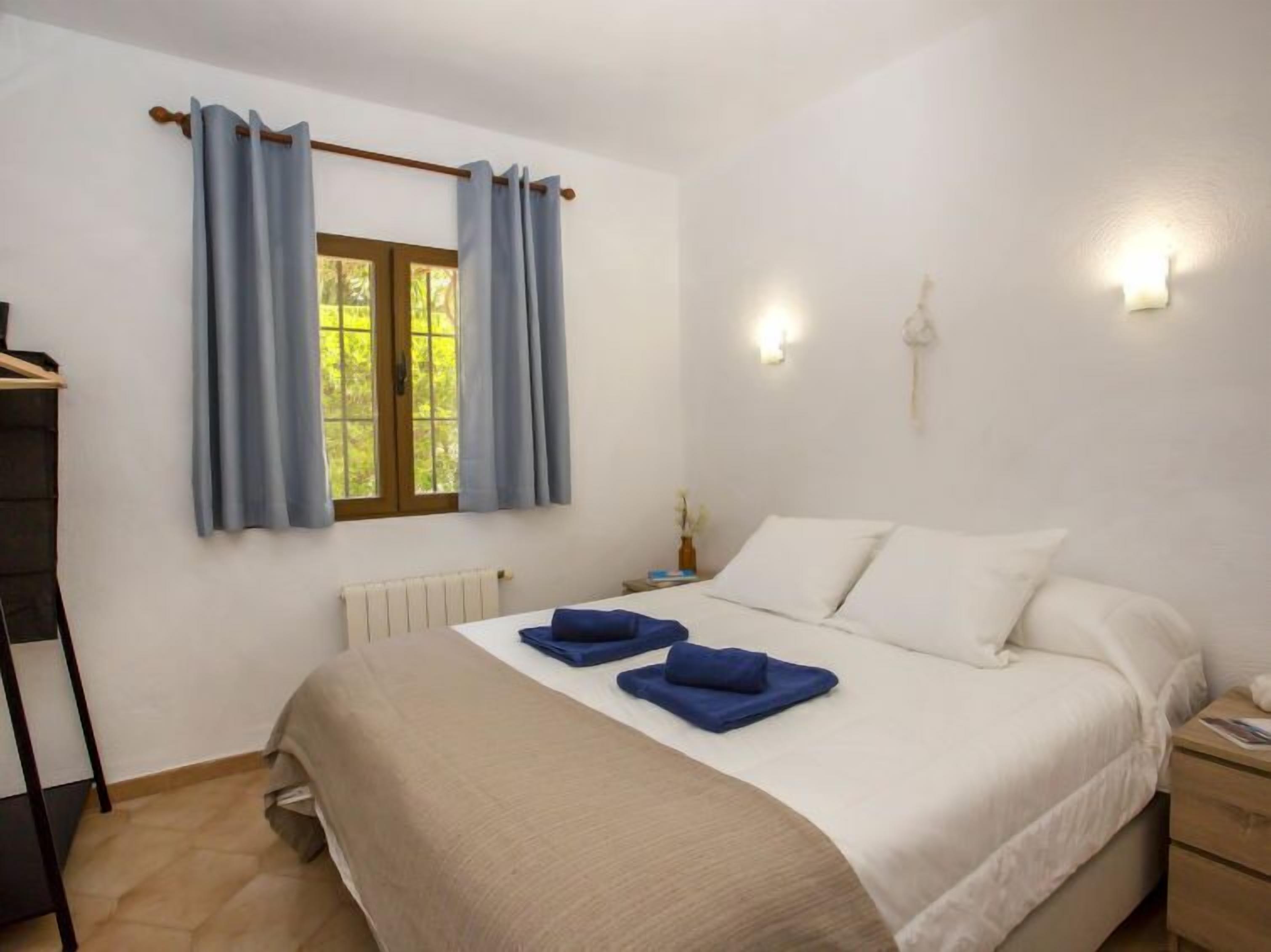 Quarto com cama, mesa de cabeceira, janela com cortinas e luminária na parede.