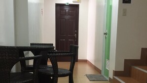 Un pasillo con una zona de comedor que incluye sillas de mimbre y una mesa, una puerta verde y un letrero de precaución sobre un piso resbaladizo.