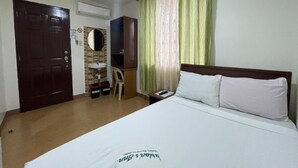 Una habitación de hotel con cama, una puerta de madera, un escritorio pequeño con silla, un espejo y una ventana con cortinas.