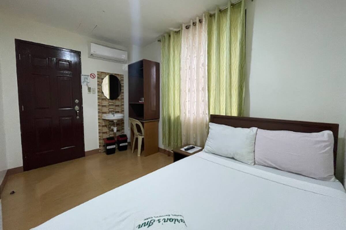 A hotel room with a bed, a wooden door, a small desk with a chair, a mirror, and a window with curtains.