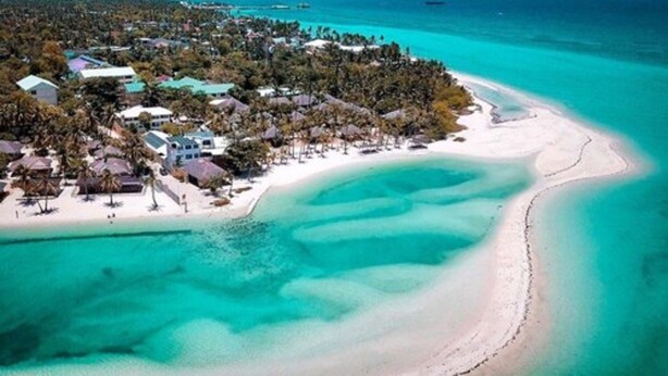 Un complejo turístico frente al mar con una playa de arena blanca, aguas turquesas cristalinas y un denso conjunto de edificios con techos de paja.