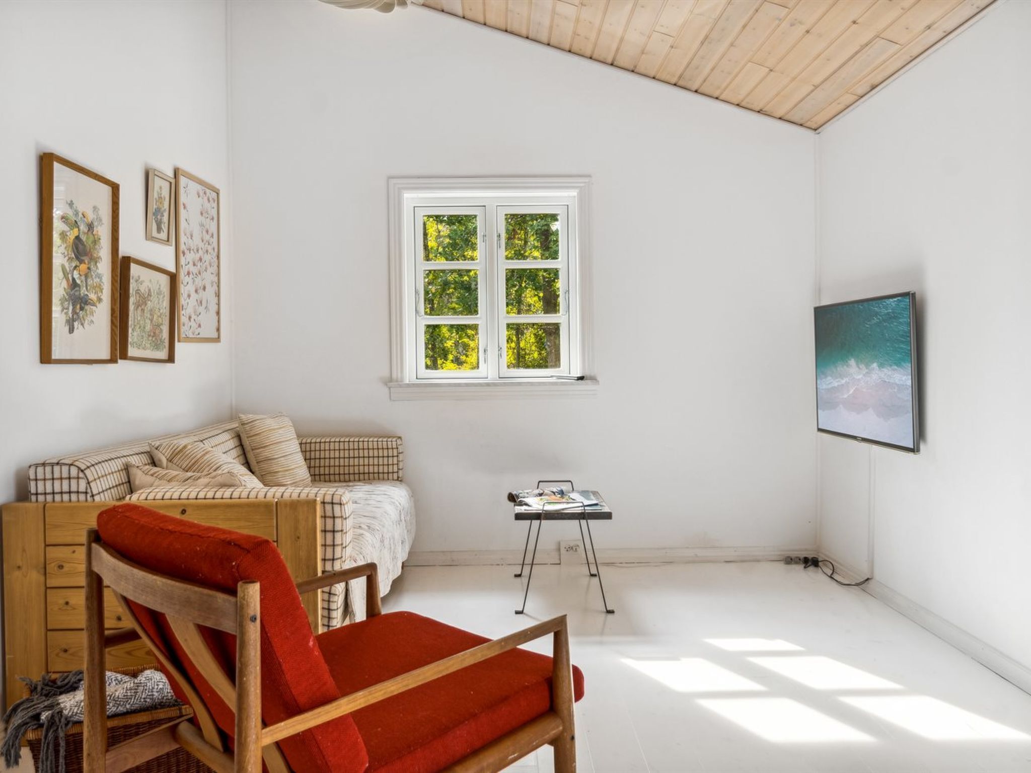Une pièce avec un plafond en bois, une fenêtre donnant sur les arbres, un canapé, un fauteuil et un téléviseur à écran plat fixé au mur.
