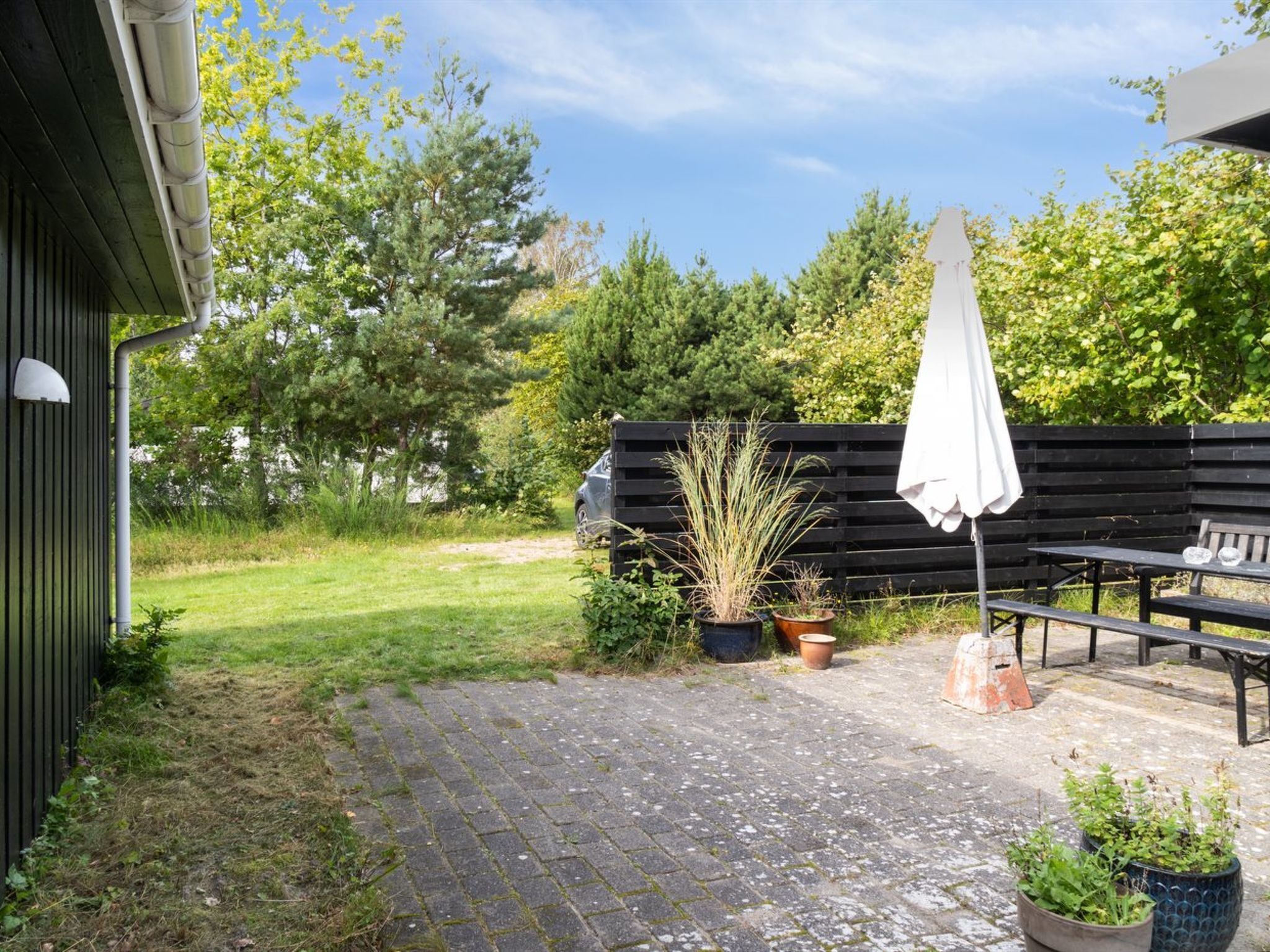 A paved patio with a bench and an umbrella, surrounded by potted plants and a wooden fence.
