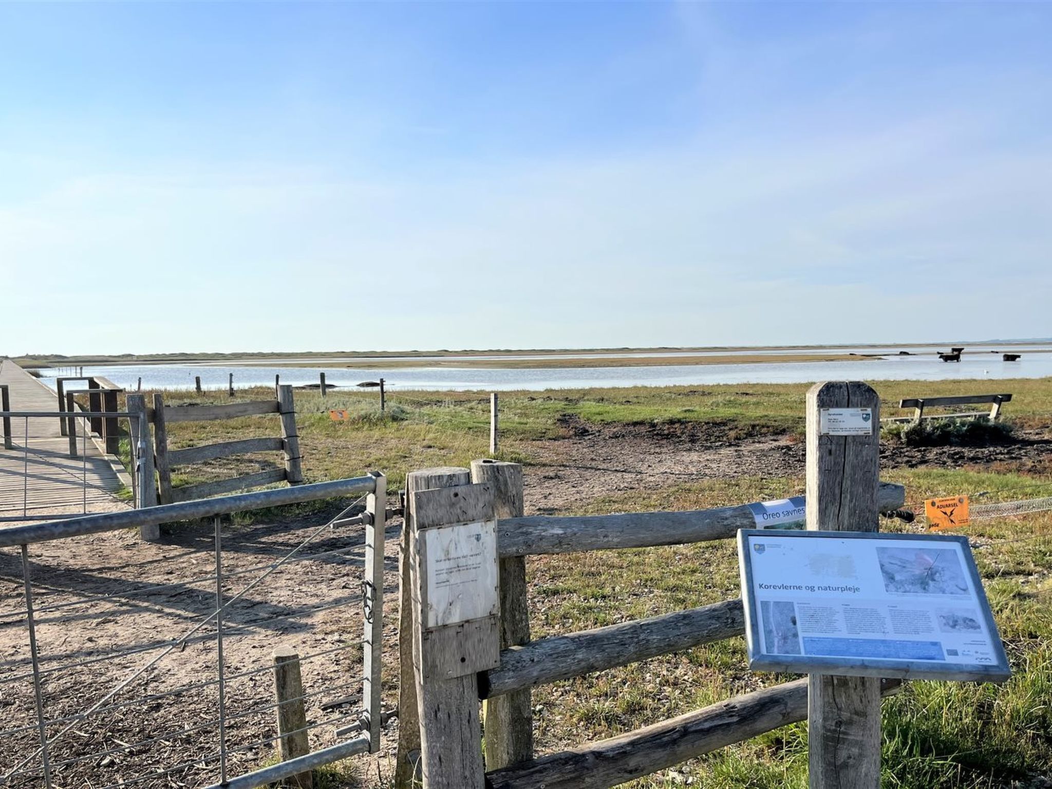 Une clôture en bois, avec un panneau indicateur et une plaque portant un texte, surplombant une étendue d’eau.