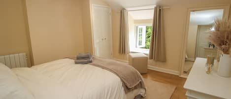A bedroom with a large bed, a bedside table with a vase, a window with curtains, and a wooden ceiling beam.