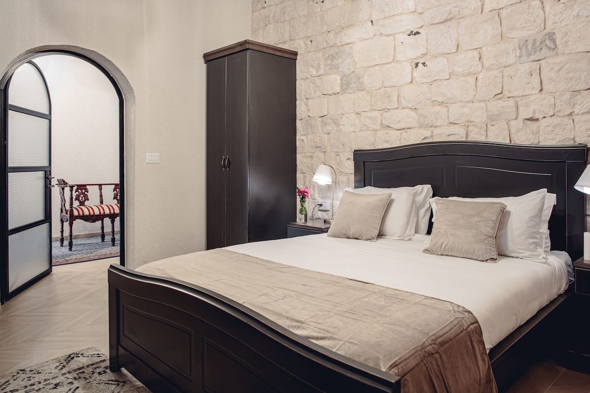 A bedroom with a stone wall, a bed with a wooden headboard, a nightstand with a lamp, and a wardrobe.