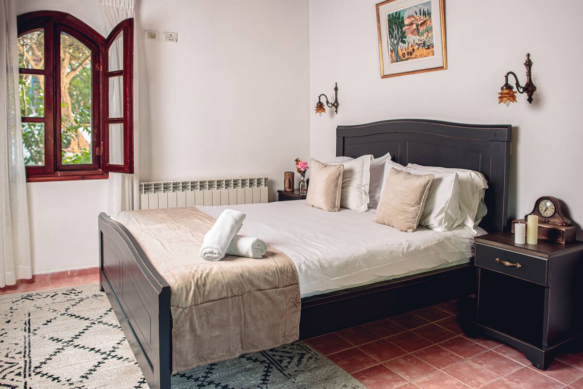 A neatly made bed with a white comforter and beige pillows, a dark wooden nightstand with a clock and candles, and a patterned rug on a tiled floor.