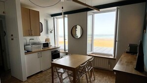 A compact kitchen with a dining table, wooden cabinets, and a large window offering a view of the beach. - À L'est - Studio vue mer sur la Digue (Arromanches-les-Bains)