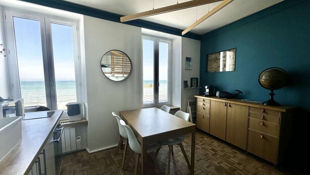A compact kitchen with a wooden table and chairs, a wooden cabinet, and a view of the sea through large windows. - À L'est - Studio vue mer sur la Digue (Arromanches-les-Bains)