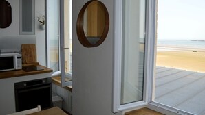 A kitchen with a microwave, wooden cutting board, and a view of the beach through a large window. - À L'est - Studio vue mer sur la Digue (Arromanches-les-Bains)