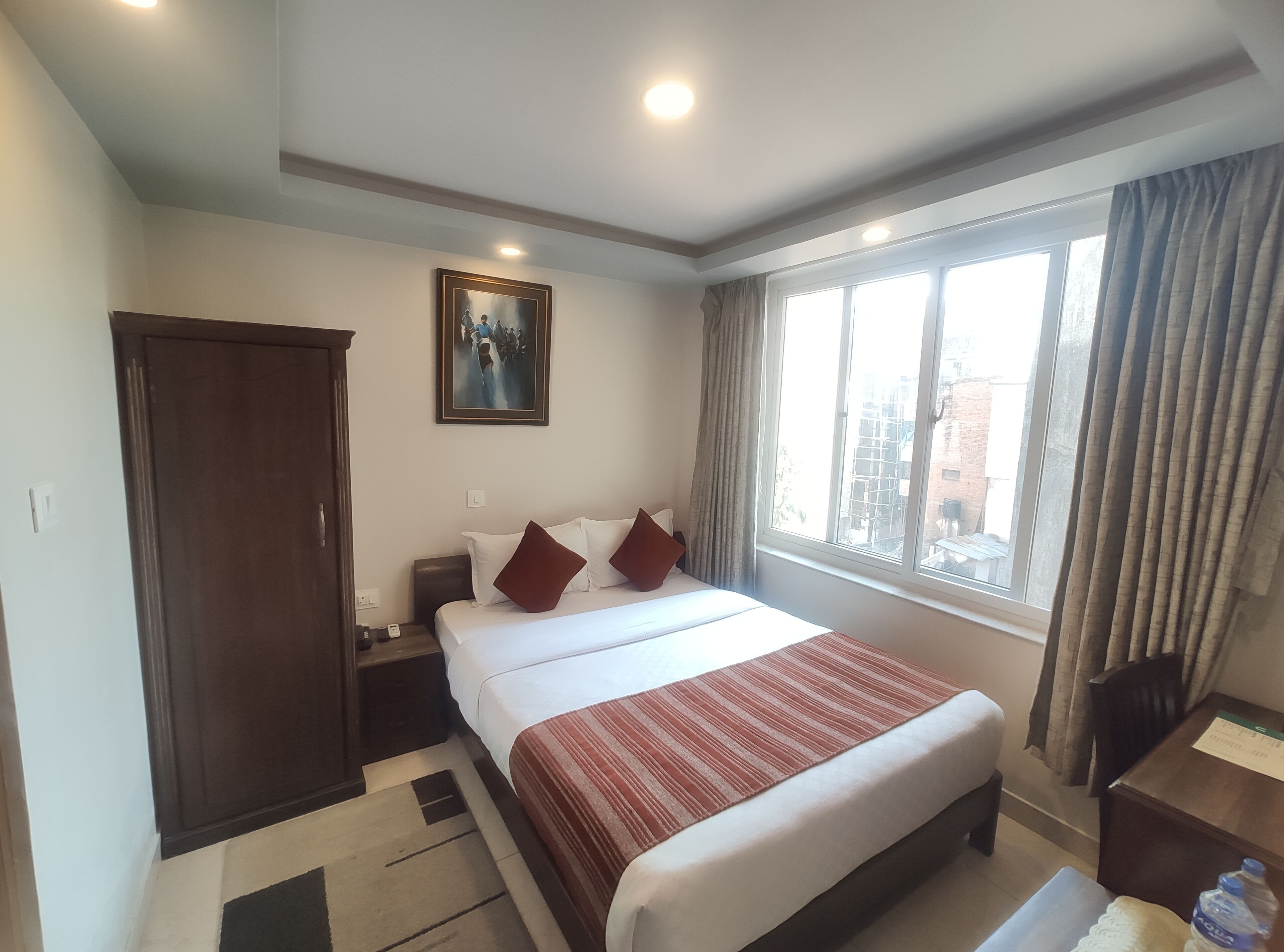 A hotel room with a bed, a nightstand, a desk, a chair, a window with curtains, a door, and a painting on the wall.