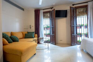 Una habitación con sofá, una mesita, cama, televisor y ventana con vista a plantas.