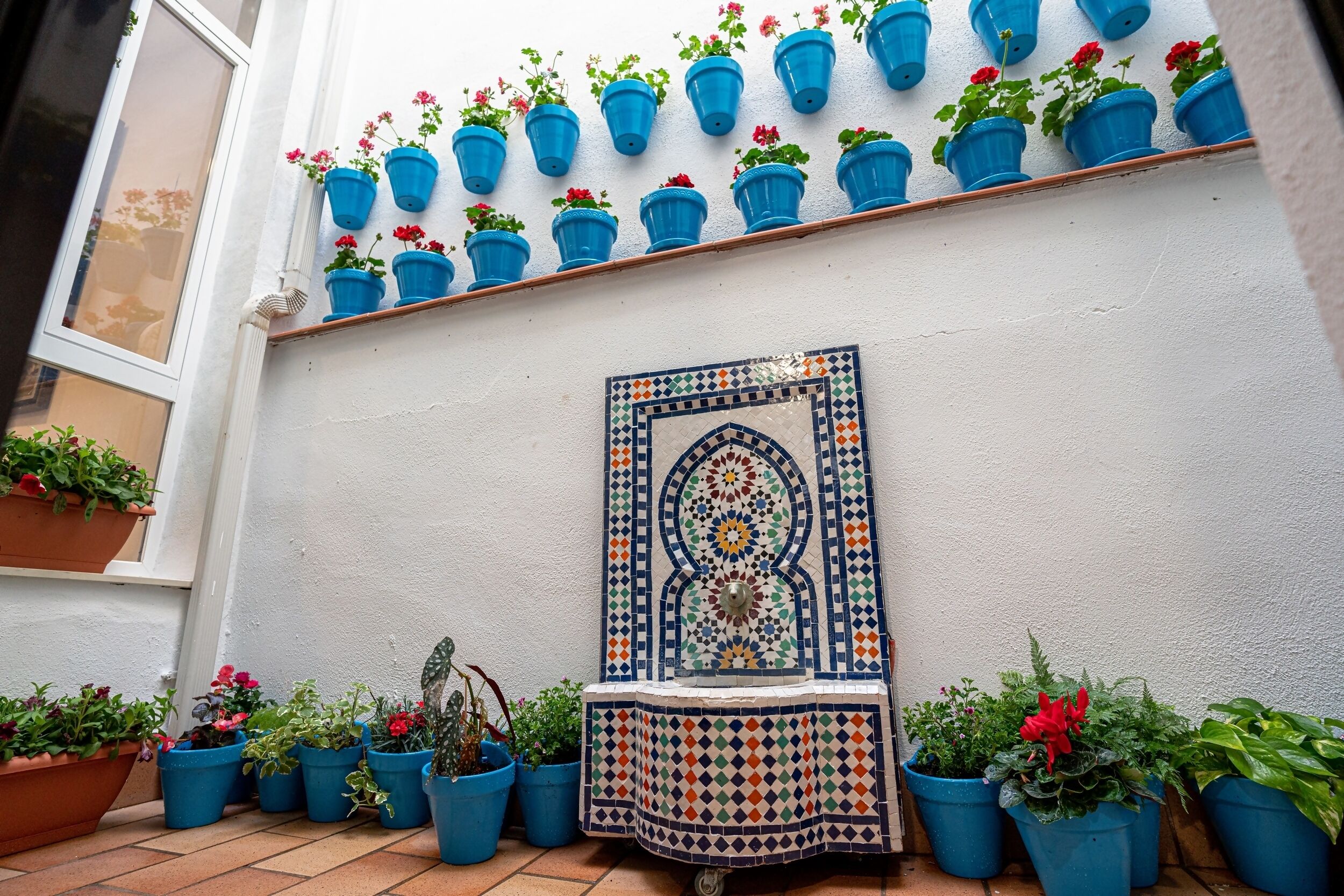 Pared de azulejos con un panel decorativo, plantas en macetas azules y una ventana.