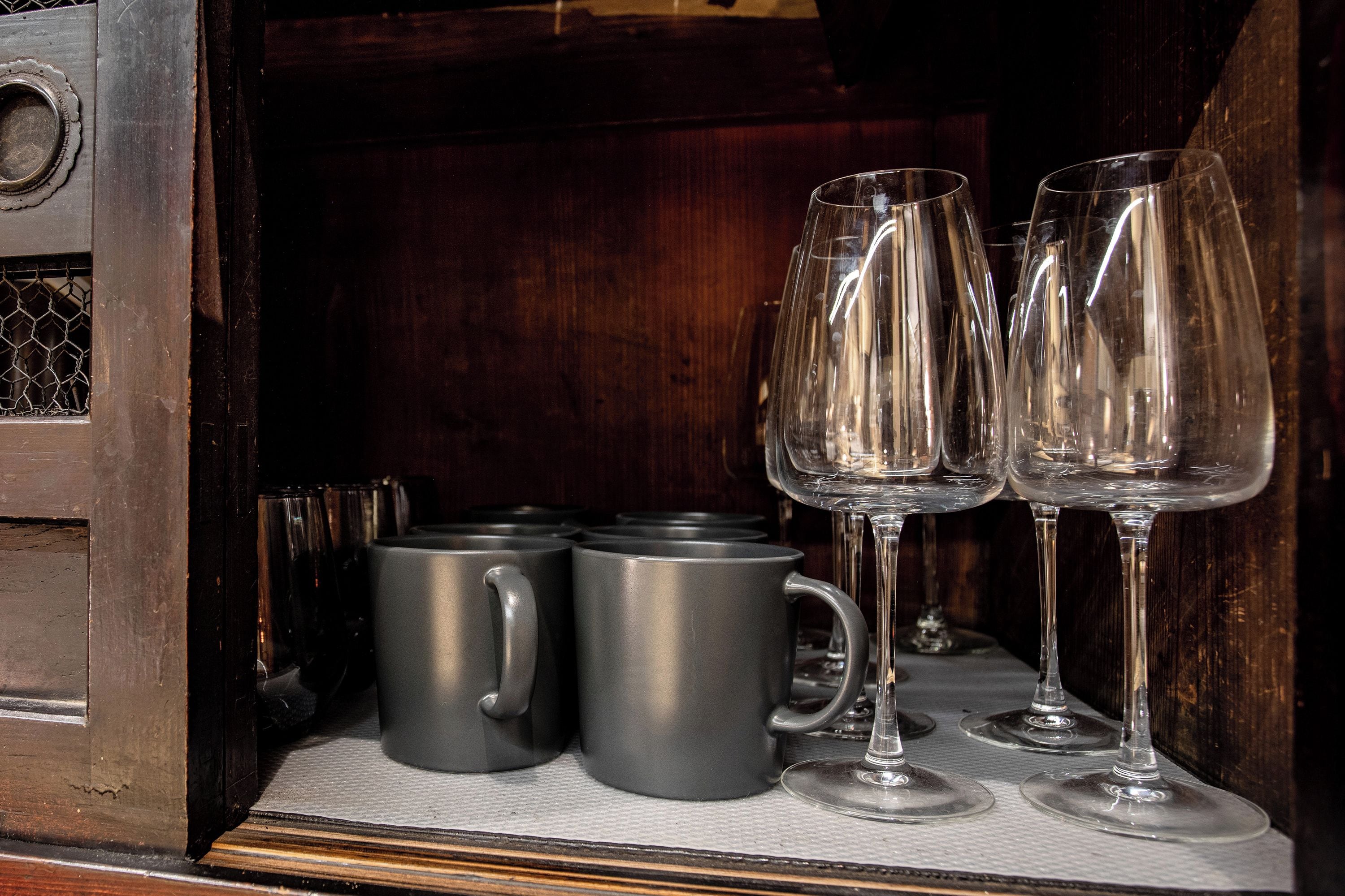 A wooden cabinet with a metal latch, containing a set of grey mugs and four wine glasses.